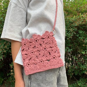 Peut inclure: Un sac à bandoulière en crochet rose poussiéreux avec un motif floral. Le sac est doté d'une bandoulière tressée et d'un anneau en métal argenté. Le sac est fait d'une matière douce et texturée et se porte à l'épaule.