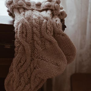 Puede incluir: Un par de calcetines de punto en un suave tono rosa apagado. Los calcetines presentan un diseño de punto trenzado con un puño texturizado con bolitas. Los calcetines acogedores están doblados sobre una superficie oscura, lo que sugiere un artículo hecho a mano.