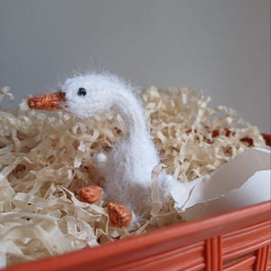 May include: A handmade white knitted goose emerging from a broken eggshell, nestled in shredded paper inside an orange woven basket. The goose has an orange beak and feet. The background is a neutral color.