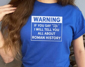 Op de afbeelding: Koningsblauw T-shirt met witte tekst: "WARNING IF YOU SAY 'Hi' I WILL TELL YOU ALL ABOUT ROMAN HISTORY". Het shirt heeft korte mouwen en een ronde hals.