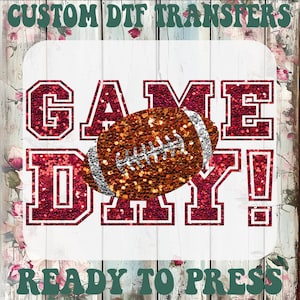 May include: A DTF transfer sheet featuring the text GAME DAY! in a dark red glitter font with a white outline. A brown and silver glitter American football graphic is centered over the text. The background has a white wood and floral pattern.