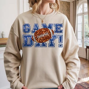 Puede incluir: Una sudadera beige de cuello redondo con el texto GAME DAY en textura de purpurina azul. Un bal&oacute;n de f&uacute;tbol americano marr&oacute;n con textura de purpurina plateada est&aacute; situado en el centro del texto. El dise&ntilde;o tiene un estilo de letras de bloque.