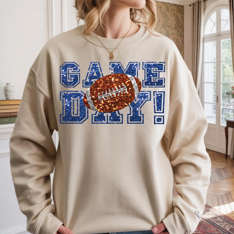 Puede incluir: Una sudadera beige de cuello redondo con el texto GAME DAY en textura de purpurina azul. Un bal&oacute;n de f&uacute;tbol americano marr&oacute;n con textura de purpurina plateada est&aacute; situado en el centro del texto. El dise&ntilde;o tiene un estilo de letras de bloque.