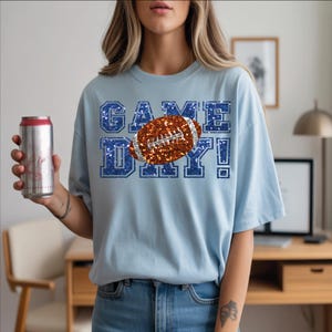 Puede incluir: Una camiseta de manga corta de color azul claro con un estampado gr&aacute;fico que muestra el texto GAME DAY en una textura de purpurina azul. Un bal&oacute;n de f&uacute;tbol americano marr&oacute;n con textura brillante y cordones plateados est&aacute; en el centro.