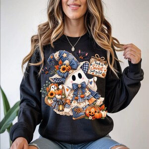 May include: A black crewneck sweatshirt featuring a graphic of a ghost wearing a blue plaid witch hat and dress. The ghost holds a sign that reads Trick or Sweet Smell my Feet, surrounded by sunflowers, pumpkins, a lantern, and small bats.
