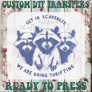 May include: A DTF transfer design featuring three raccoons in a blue and white illustration. The text reads "Get in Scavenger, We are going thrifting, Ready to press". The design is on a white background with a floral and wood grain pattern.