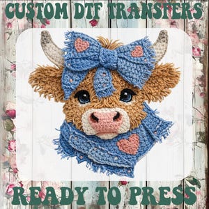 May include: A crocheted cow face with a blue bow and scarf, featuring pink hearts. The image includes the text "CUSTOM DTF TRANSFERS" and "READY TO PRESS" in a green, decorative font. The cow has brown fur, a pink nose, and blue eyes.