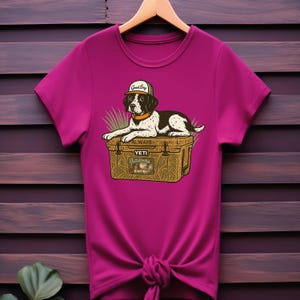 Peut inclure: T-shirt magenta avec un dessin anim&eacute; de chien portant une casquette avec l'inscription "Good Boy". Le chien est allong&eacute; sur une glaci&egrave;re avec le texte "ALWAYS YETI Outdoors Hunting". Le t-shirt est nou&eacute; &agrave; la taille et est suspendu &agrave; un cintre en bois.