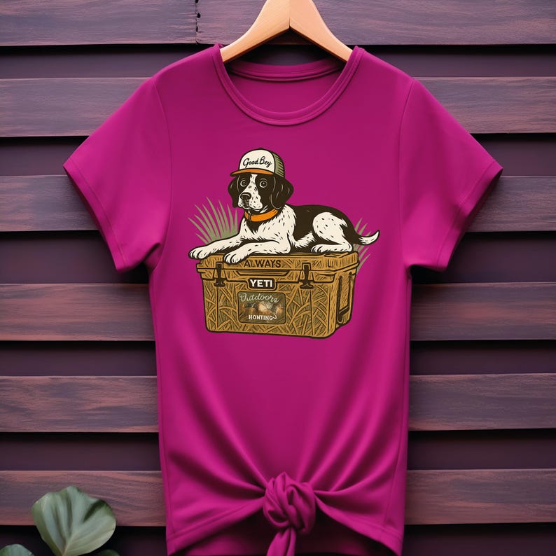 Peut inclure: T-shirt magenta avec un dessin anim&eacute; de chien portant une casquette avec l'inscription "Good Boy". Le chien est allong&eacute; sur une glaci&egrave;re avec le texte "ALWAYS YETI Outdoors Hunting". Le t-shirt est nou&eacute; &agrave; la taille et est suspendu &agrave; un cintre en bois.