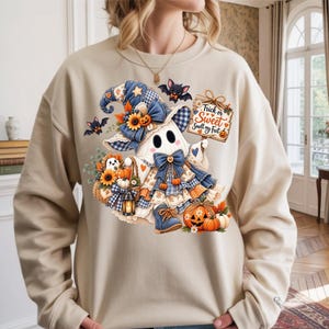 May include: A beige crewneck sweatshirt featuring a graphic of a ghost wearing a blue plaid witch hat and dress, holding a sign that reads Trick or Sweet Smell my Feet. The design includes sunflowers, pumpkins, bats, and a lantern.