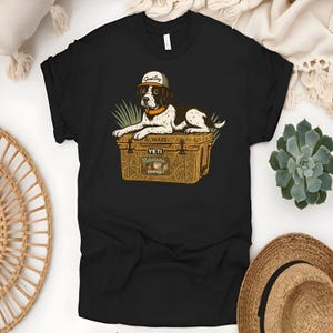 Peut inclure: T-shirt noir avec un dessin d'un chien portant une casquette avec l'inscription "Good Boy" et reposant sur une glaci&egrave;re. La glaci&egrave;re porte l'inscription "ALWAYS YETI Outdoors Hunting". Le chien est marron, noir et blanc.