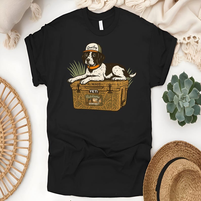 Peut inclure: T-shirt noir avec un dessin d'un chien portant une casquette avec l'inscription "Good Boy" et reposant sur une glaci&egrave;re. La glaci&egrave;re porte l'inscription "ALWAYS YETI Outdoors Hunting". Le chien est marron, noir et blanc.
