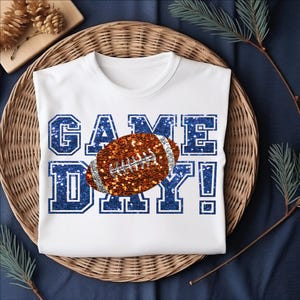 Puede incluir: Una camiseta blanca de manga corta con el texto GAME DAY en una fuente de estilo purpurina azul. Un gr&aacute;fico de un bal&oacute;n de f&uacute;tbol americano de estilo purpurina marr&oacute;n se sit&uacute;a en el centro, superponi&eacute;ndose a las letras con cordones plateados.
