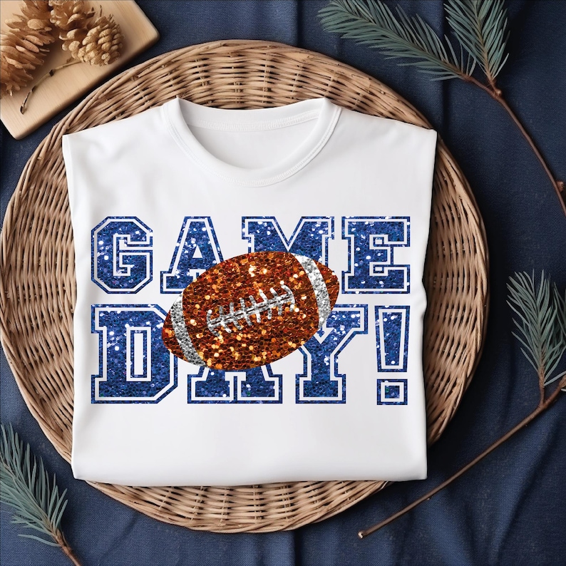 Puede incluir: Una camiseta blanca de manga corta con el texto GAME DAY en una fuente de estilo purpurina azul. Un gr&aacute;fico de un bal&oacute;n de f&uacute;tbol americano de estilo purpurina marr&oacute;n se sit&uacute;a en el centro, superponi&eacute;ndose a las letras con cordones plateados.