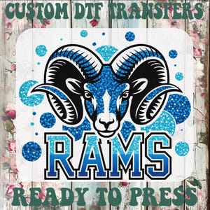May include: A DTF transfer design featuring a ram's head with large black horns and a blue glitter design. The word "RAMS" is in blue and white block letters. The text "CUSTOM DTF TRANSFERS" and "READY TO PRESS" are at the top and bottom.