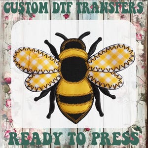 May include: A detailed, embroidered bee design with yellow and black coloring. The bee's wings feature a yellow and white gingham pattern. The text "CUSTOM DTF TRANSFERS" and "READY TO PRESS" are displayed in green, arched across the top and bottom of the image.