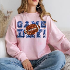 Puede incluir: Una sudadera de cuello redondo rosa claro con un estampado gr&aacute;fico que muestra el texto GAME DAY en una textura azul brillante. Un bal&oacute;n de f&uacute;tbol americano marr&oacute;n con una textura plateada brillante est&aacute; colocado en el centro del texto.