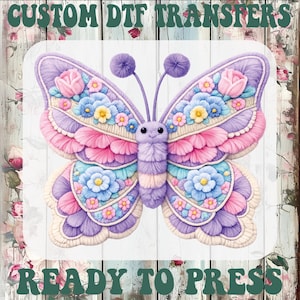 May include: A colorful butterfly design with pink, blue, and purple accents, adorned with floral details. The image includes the text "CUSTOM DTF TRANSFERS" and "READY TO PRESS" in a green, vintage font. The butterfly is centered on a white wooden background.