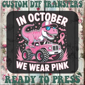 May include: A custom DTF transfer featuring a pink dinosaur wearing a baseball cap and sunglasses, standing on a pink monster truck. The design includes a pink awareness ribbon, stars, and the text "IN OCTOBER WE WEAR PINK" on a dark background.
