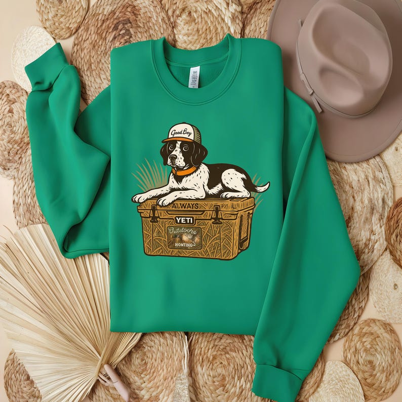 Peut inclure: Un sweat-shirt vert avec un dessin d'un chien portant une casquette avec l'inscription "Good Boy" et reposant sur une glaci&egrave;re. La glaci&egrave;re porte l'inscription "Yeti Outdoors Hunting". Le sweat-shirt est pli&eacute; sur une surface tiss&eacute;e.
