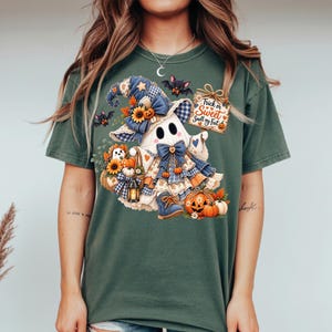 May include: A dark green t-shirt featuring a graphic of a ghost wearing a blue plaid hat and bow, holding a basket with sunflowers and pumpkins. The design includes small bats, a lantern, and a sign that reads Trick or Sweet Smell my Feet.