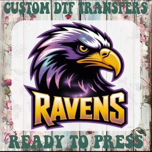 May include: A graphic featuring a stylized eagle head with the word "RAVENS" in gold lettering. The eagle has purple and black feathers, a yellow beak, and a cosmic design. The text "CUSTOM DTF TRANSFERS" and "READY TO PRESS" are at the top and bottom.