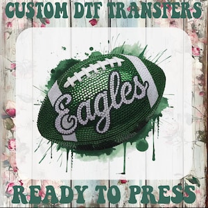 May include: A green football with the word "Eagles" in a sparkling font, set against a green watercolor splash. The image includes the text "CUSTOM DTF TRANSFERS" and "READY TO PRESS" in a decorative font, all on a white wood-grain background.