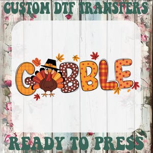 May include: A custom DTF transfer featuring the word GOBBLE in orange and brown patterns, with a cartoon turkey wearing a black pilgrim hat replacing the letter O. The design includes small autumn leaves and the text CUSTOM DTF TRANSFERS READY TO PRESS.