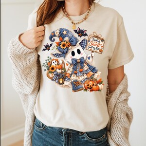 May include: A beige t-shirt featuring a graphic of a ghost wearing a blue plaid hat and dress, holding a sign that reads Trick or Sweet Smell my Feet. The design includes sunflowers, pumpkins, bats, and a lantern, all in a colorful illustration.