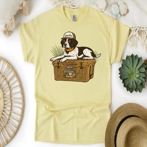 Peut inclure: T-shirt jaune clair avec un dessin d'un chien portant une casquette avec l'inscription "Good Boy" et reposant sur une glaci&egrave;re marron. La glaci&egrave;re porte l'inscription "Yeti Outdoors Hunting" et le mot "Always".