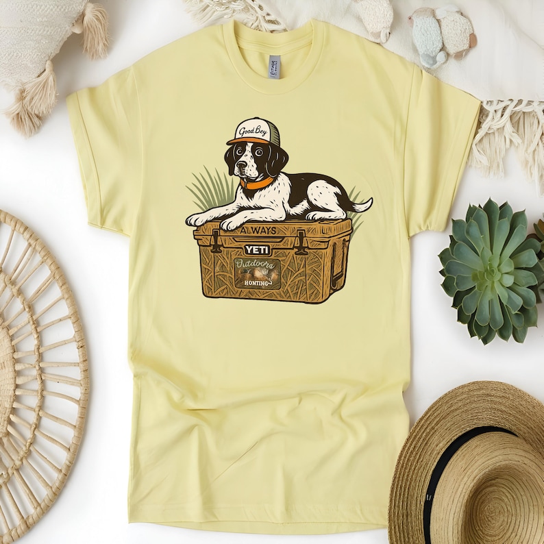 Peut inclure: T-shirt jaune clair avec un dessin d'un chien portant une casquette avec l'inscription "Good Boy" et reposant sur une glaci&egrave;re marron. La glaci&egrave;re porte l'inscription "Yeti Outdoors Hunting" et le mot "Always".