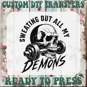 May include: Black and white graphic of a skull holding a barbell, with the text "Sweating Out All My Demons" and "Ready to Press". The design is for DTF transfers.