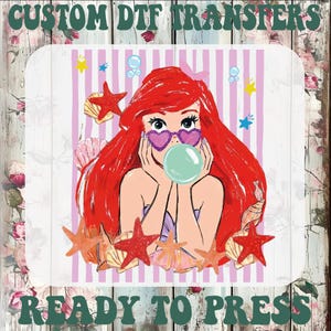 May include: DTF transfer featuring a cartoon illustration of a mermaid with red hair, heart-shaped sunglasses, and a bubble. The design includes starfish, seashells, and the text "CUSTOM DTF TRANSFERS" and "READY TO PRESS".