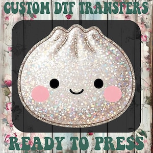 May include: A custom DTF transfer featuring a holographic, glittery dumpling character with a smiling face, black circular eyes, and pink circular cheeks. The text CUSTOM DTF TRANSFERS and READY TO PRESS is displayed in dark green lettering.