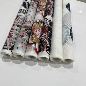 Peut inclure: Plusieurs papiers d&eacute;coratifs roul&eacute;s avec divers motifs. Les motifs comprennent du texte, des bonhommes en pain d'&eacute;pices festifs et des motifs sportifs. Les papiers sont blancs avec des motifs et du texte color&eacute;s, parfaits pour l'artisanat et les d&eacute;corations.