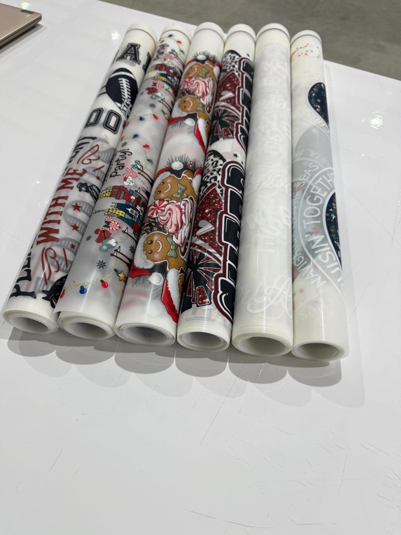 Peut inclure: Plusieurs papiers d&eacute;coratifs roul&eacute;s avec divers motifs. Les motifs comprennent du texte, des bonhommes en pain d'&eacute;pices festifs et des motifs sportifs. Les papiers sont blancs avec des motifs et du texte color&eacute;s, parfaits pour l'artisanat et les d&eacute;corations.