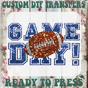 Puede incluir: Una transferencia DTF personalizada con el texto GAME DAY! en letras de bloque azul brillante. Un bal&oacute;n de f&uacute;tbol americano marr&oacute;n brillante con cordones plateados est&aacute; centrado sobre el texto, sobre un fondo blanco con flores.
