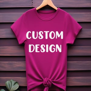May include: A pink t-shirt with short sleeves and a knotted bottom. The shirt has the text "CUSTOM DESIGN" printed in white on the front.