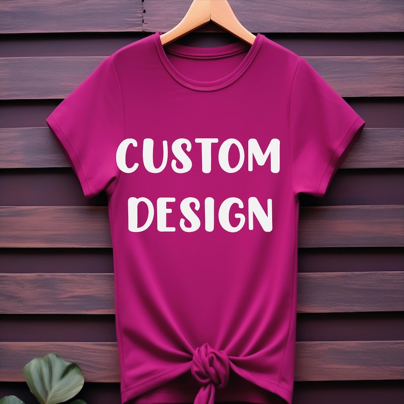 May include: A pink t-shirt with short sleeves and a knotted bottom. The shirt has the text "CUSTOM DESIGN" printed in white on the front.