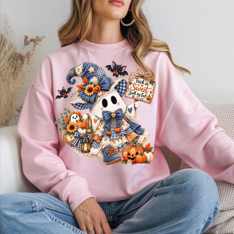 May include: A light pink sweatshirt featuring a graphic of a ghost wearing a blue plaid witch hat and boots, holding a sign that reads Trick or Sweet Smell my Feet, surrounded by pumpkins, sunflowers, and small bats in a colorful illustration.