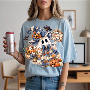 May include: A light blue t-shirt featuring a graphic of a ghost wearing a blue plaid hat and boots, holding a sign that reads Trick or Sweet Smell my Feet. The design includes sunflowers, pumpkins, a lantern, and two small bats.
