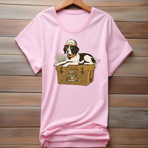 Peut inclure: T-shirt rose clair avec un dessin d'un chien noir et blanc portant une casquette avec l'inscription "Good Boy". Le chien est allong&eacute; sur une glaci&egrave;re marron avec le texte "Yeti Outdoors Hunting."