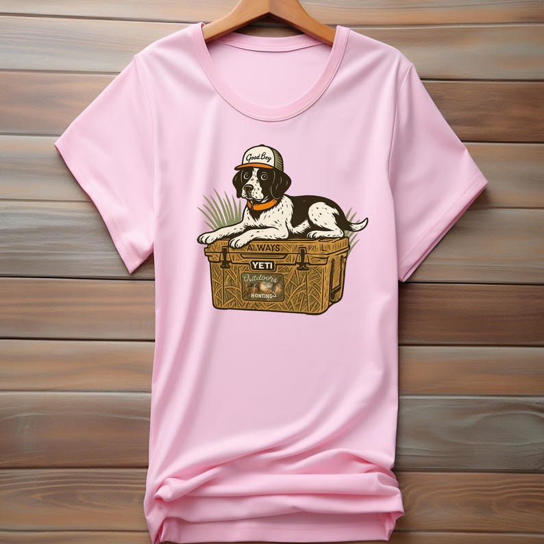 Peut inclure: T-shirt rose clair avec un dessin d'un chien noir et blanc portant une casquette avec l'inscription "Good Boy". Le chien est allong&eacute; sur une glaci&egrave;re marron avec le texte "Yeti Outdoors Hunting."