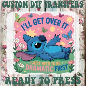 May include: A DTF transfer with a cartoon character in blue, with large eyes, lounging on grass. The design includes the text "I'll Get Over It" and "I Just Need To Be Dramatic First", surrounded by butterflies and flowers. The words "CUSTOM DTF TRANSFERS" and "READY TO PRESS" are also included.