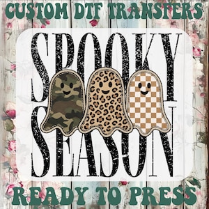 May include: A custom DTF transfer design featuring the text SPOOKY SEASON in a distressed black font. Three cartoon ghosts are centered, each with a different pattern: camouflage, leopard print, and a tan and white checkerboard pattern.