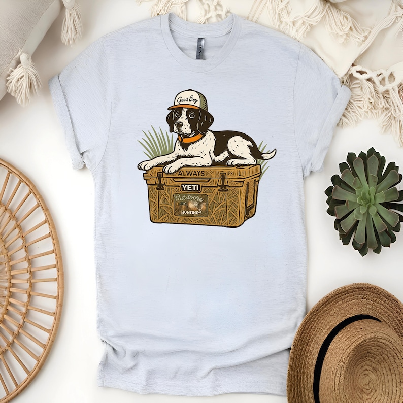 Peut inclure: T-shirt bleu clair avec un dessin anim&eacute; de chien portant une casquette avec l'inscription "Good Boy" et reposant sur une glaci&egrave;re. La glaci&egrave;re porte les inscriptions "ALWAYS" et "YETI Outdoors Hunting". Le chien est noir et blanc avec un collier orange.