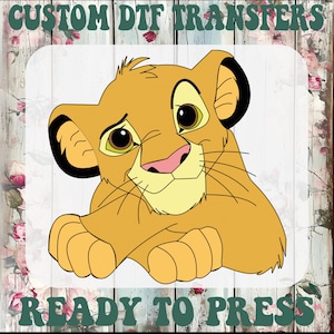 May include: A cartoon image of a lion cub with a yellow body, black ears, and a pink nose. The text "CUSTOM DTF TRANSFERS" and "READY TO PRESS" are printed above and below the lion cub image, respectively.