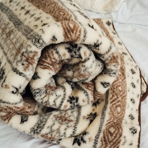 May include: Rolled-up, patterned blanket in cream, brown, and black. The blanket features a repeating geometric design with stripes and diamond shapes. The edges are trimmed with a dark brown border. The blanket is made of a soft, plush material.