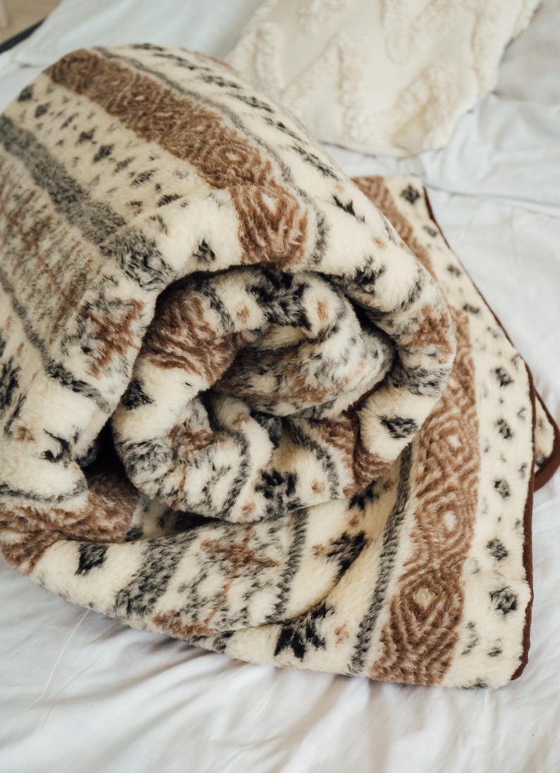 May include: Rolled-up, patterned blanket in cream, brown, and black. The blanket features a repeating geometric design with stripes and diamond shapes. The edges are trimmed with a dark brown border. The blanket is made of a soft, plush material.