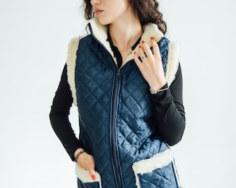May include: A quilted navy blue vest with a cream-colored faux fur trim. The vest has a zip-up front, two front pockets, and a diamond pattern. The model is wearing a black long-sleeve shirt and light blue jeans.